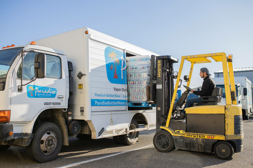 A Paradise Bottled Water truck being loaded by a forklift with various packages of Bottled Water