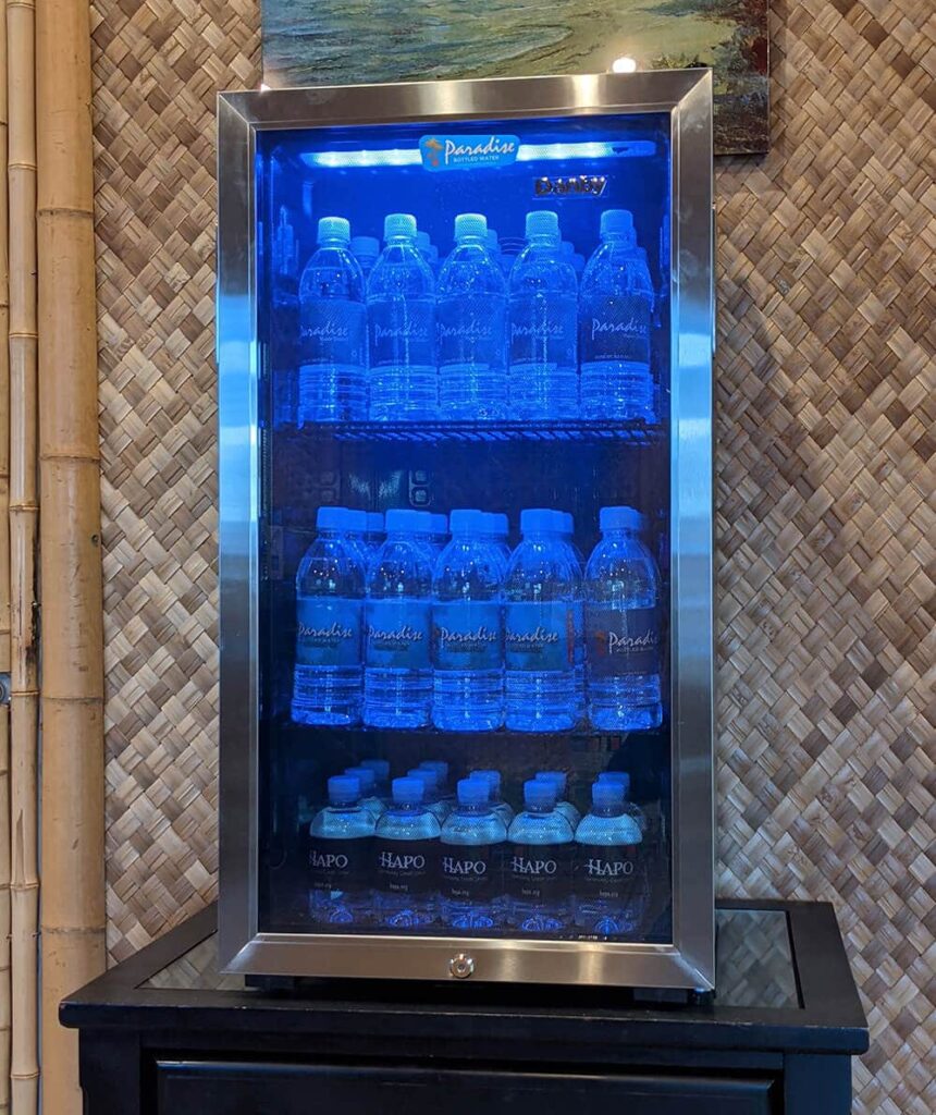 A mini-fridge with a glass door and a blue fluorescent light. The fridge is full of Paradise Water Bottles